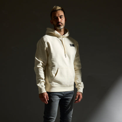 Man wearing a light-colored hoodie and dark jeans standing against a dark background